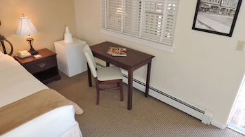 A hotel room with a bed, a desk, a chair, a lamp, a nightstand, a window with blinds, and a framed picture on the wall.