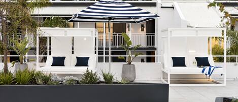A modern outdoor seating area with white sofas and a striped umbrella, surrounded by plants and a white building in the background.