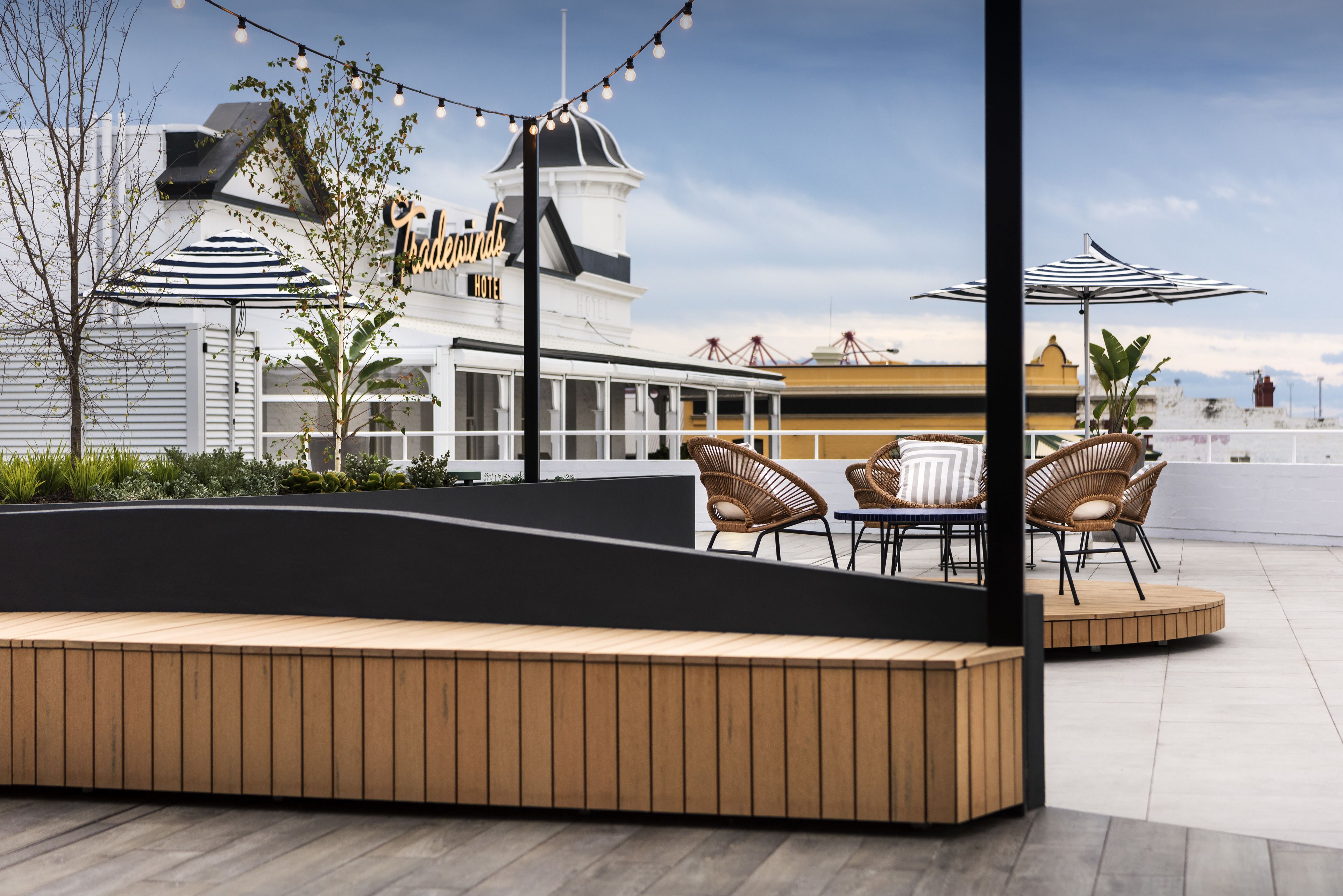 An outdoor seating area with wooden benches and wicker chairs, a white building with a sign, and string lights.