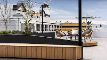 An outdoor seating area with wooden benches and wicker chairs, a white building with a sign, and string lights.