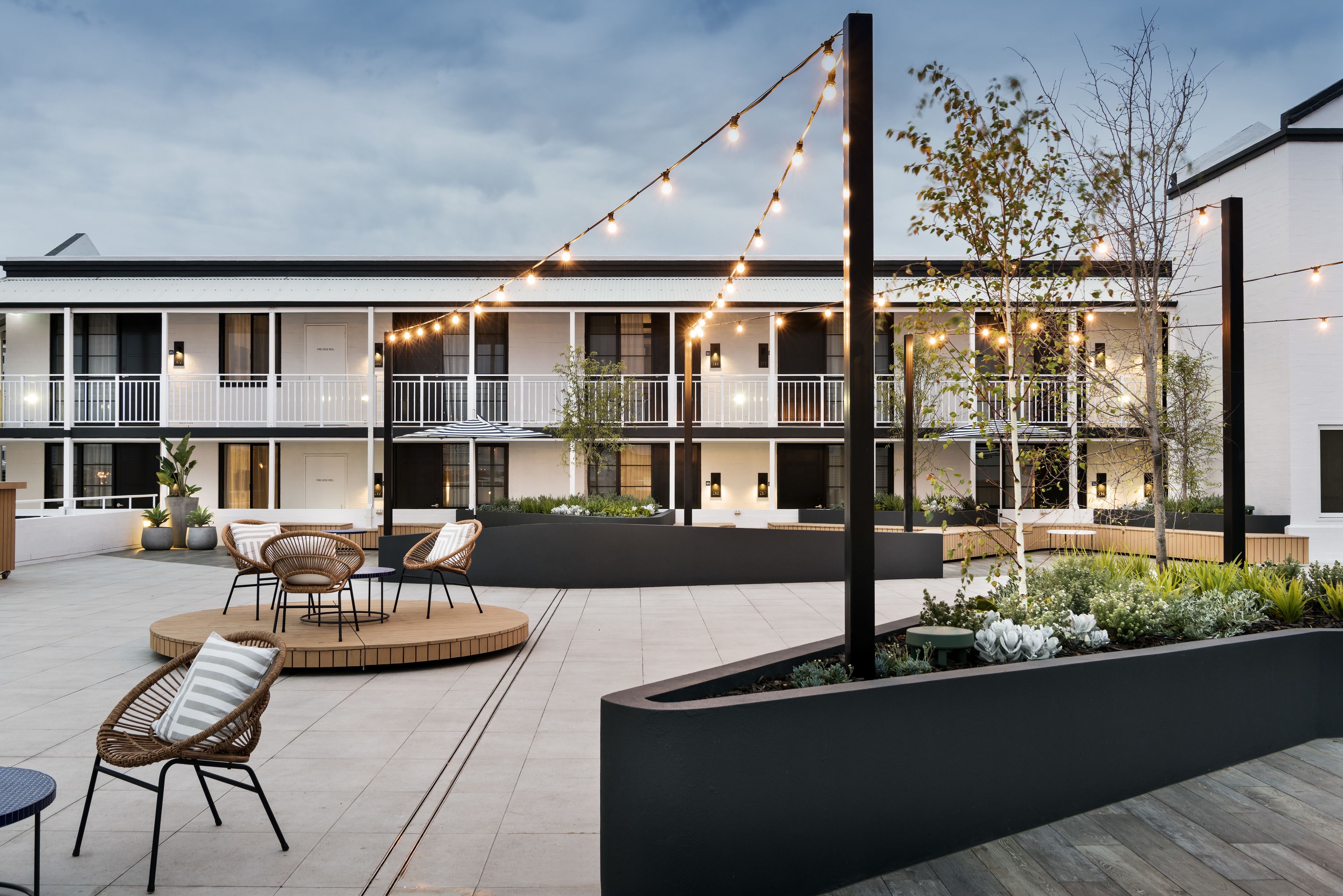 Un patio con mesas al aire libre, luces colgantes y un edificio con balcones.