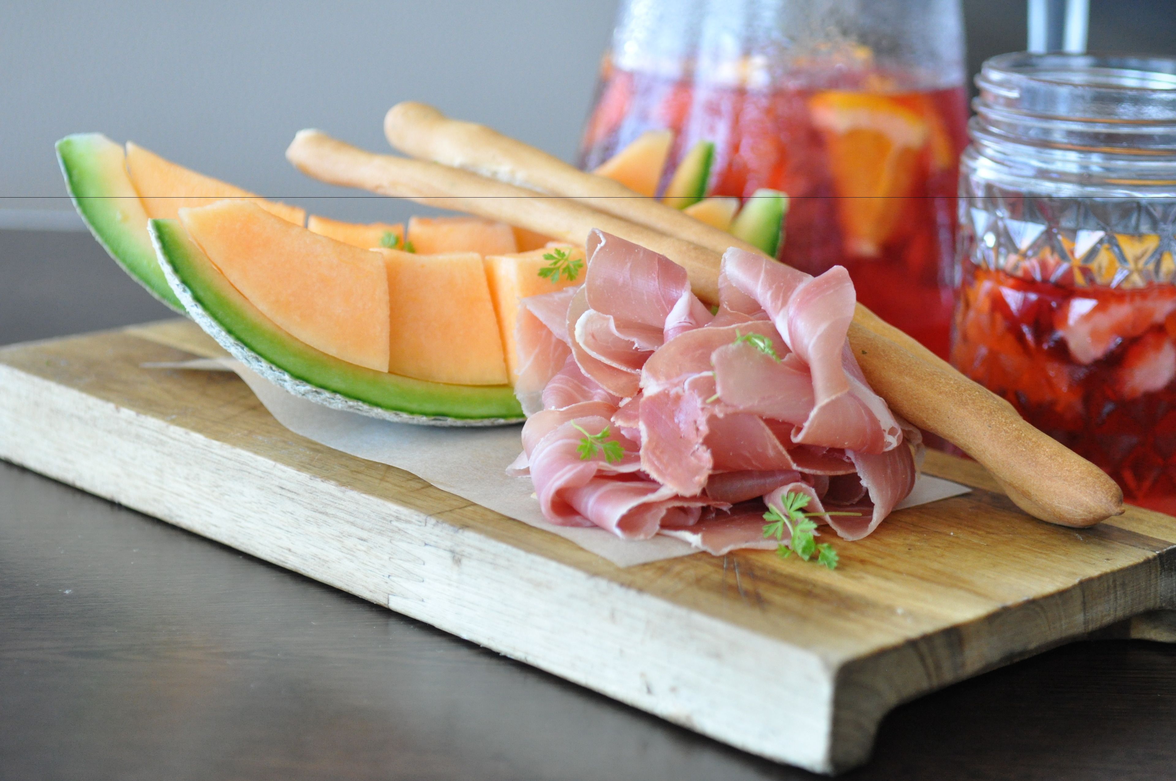 Una tabla de madera con melón en rodajas, prosciutto y un frasco con líquido rojo.