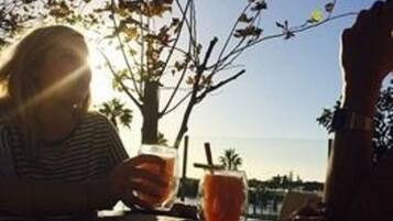 Una persona sentada en una mesa al aire libre con una bebida, luz solar y un árbol de fondo.