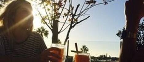A person sitting at an outdoor table with a drink, sunlight, and a tree in the background.