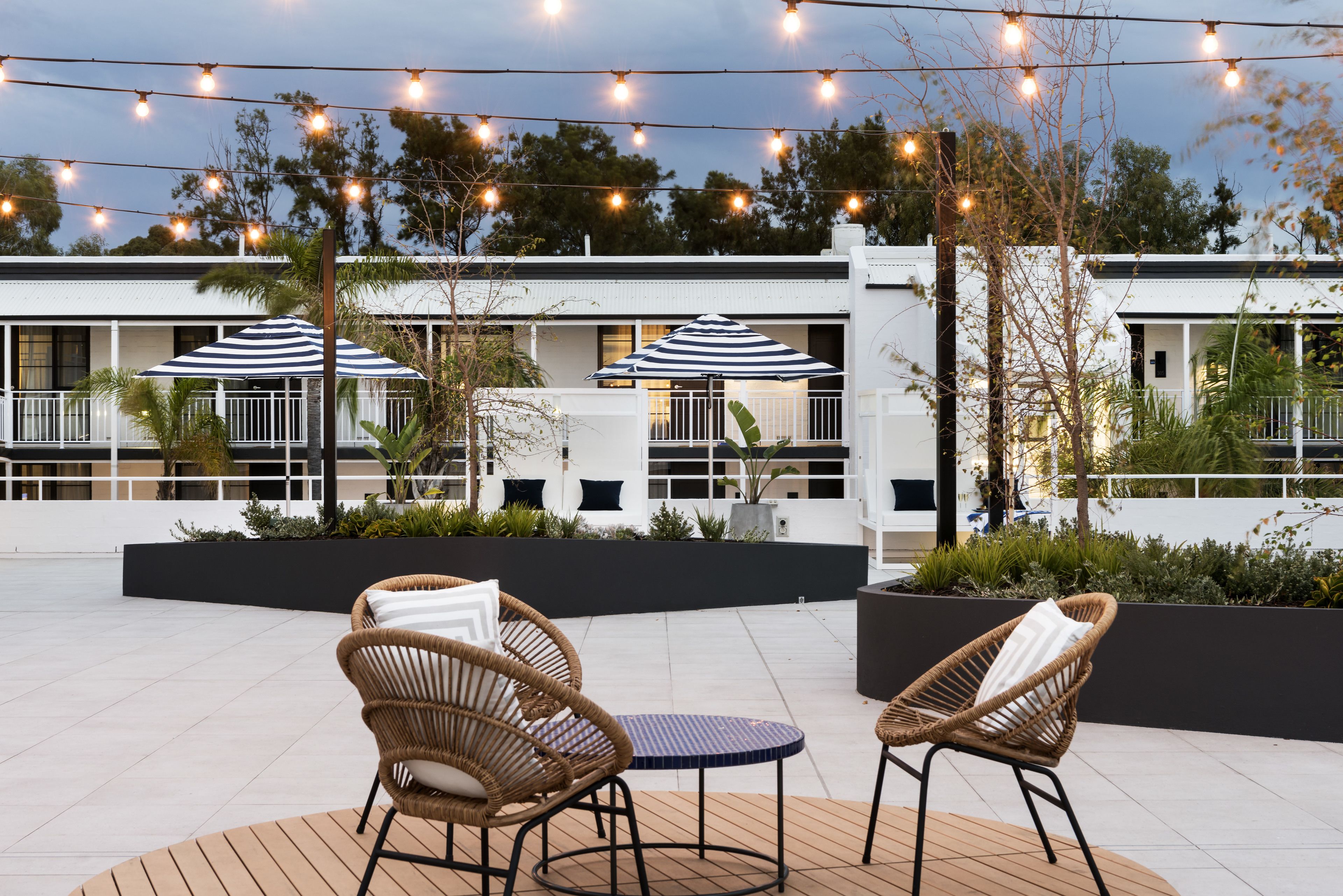 Un patio con sillas de mimbre y una mesa redonda, luces colgantes y un edificio de fachada blanca al fondo.