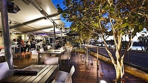 An outdoor dining area with tables and chairs, a tree, and a view of the water. - Tradewinds Hotel Fremantle (East Fremantle)
