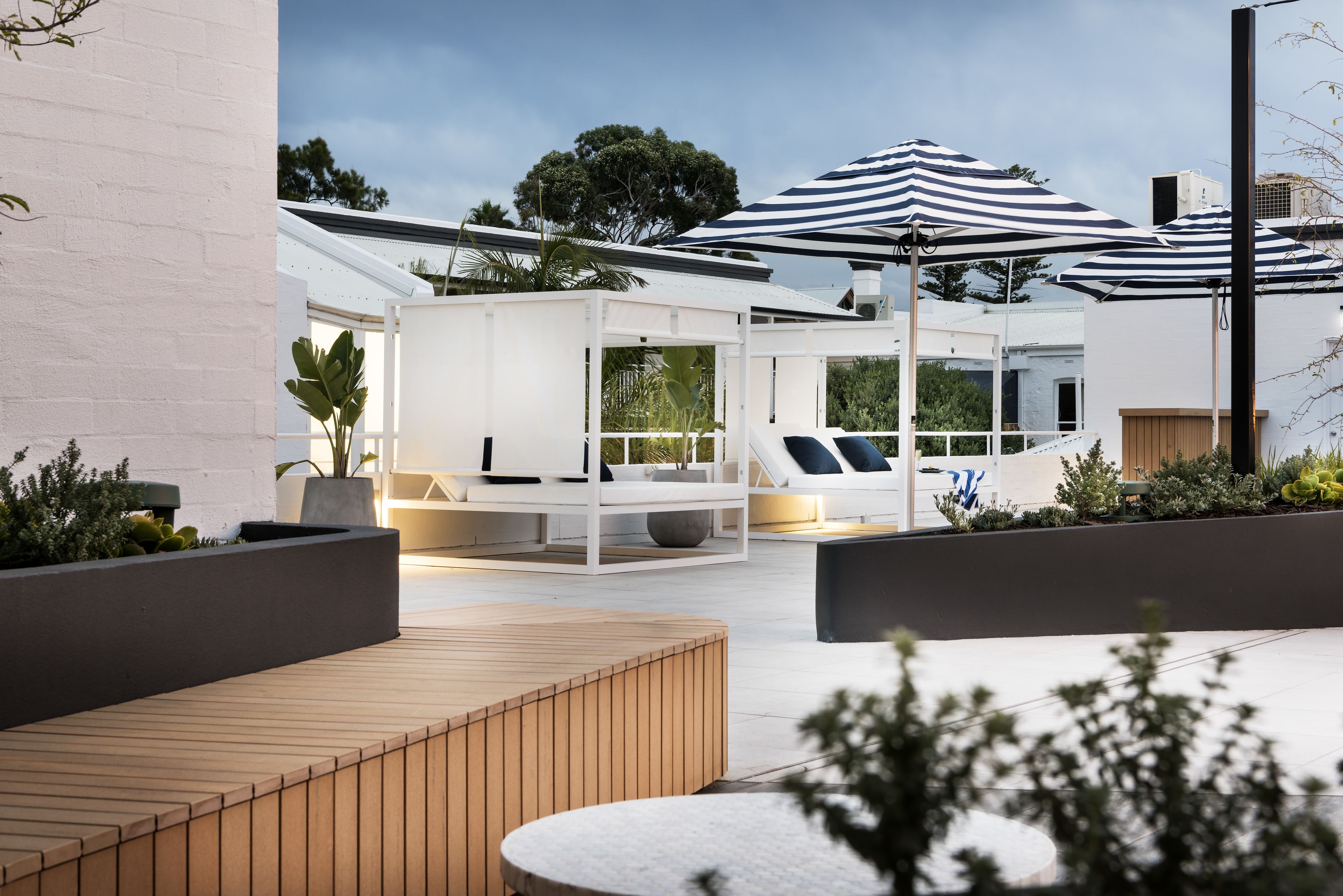 An outdoor seating area with wooden benches and tables, surrounded by planters and umbrellas.