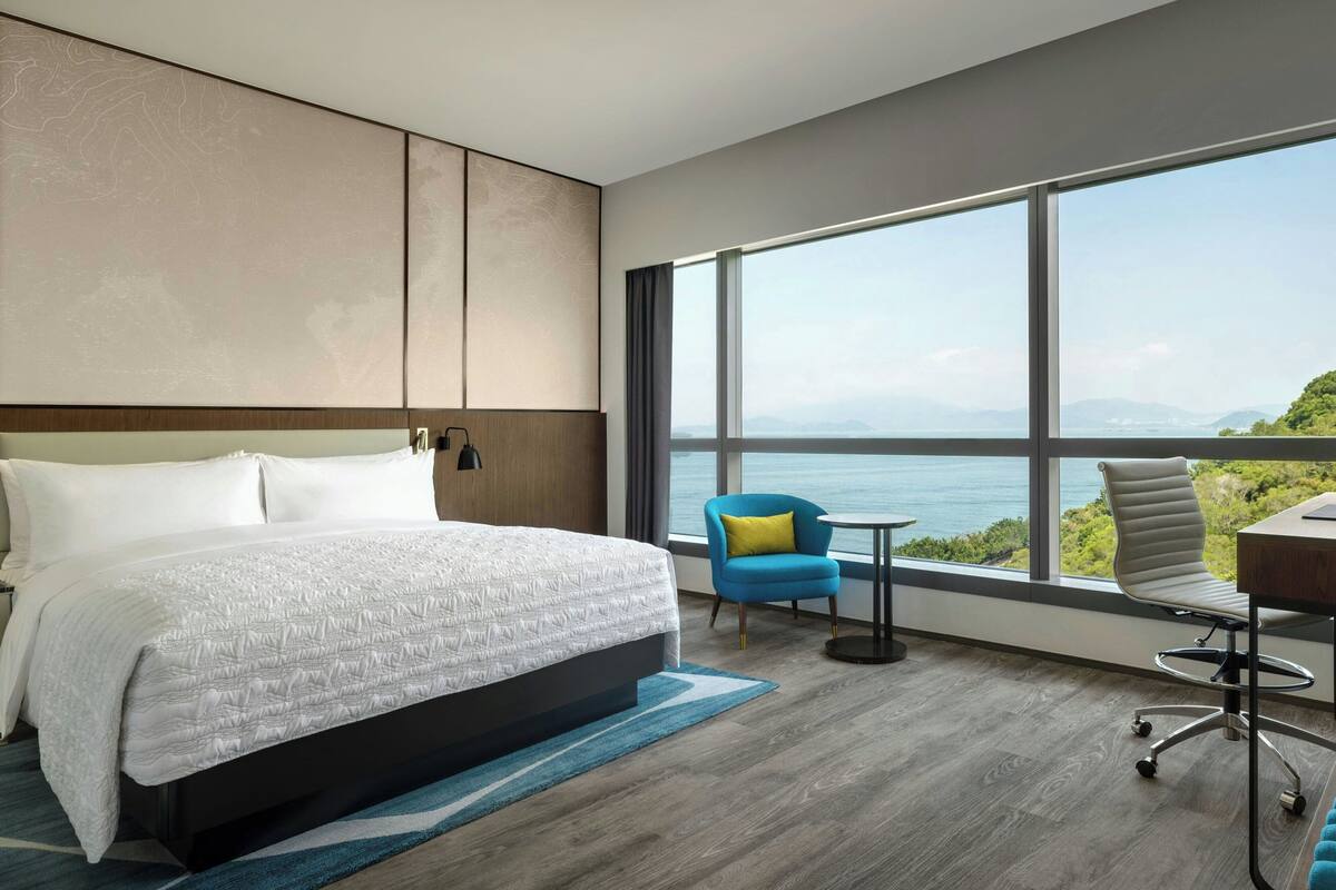 A modern hotel room with a large bed, a desk, and a view of the sea.