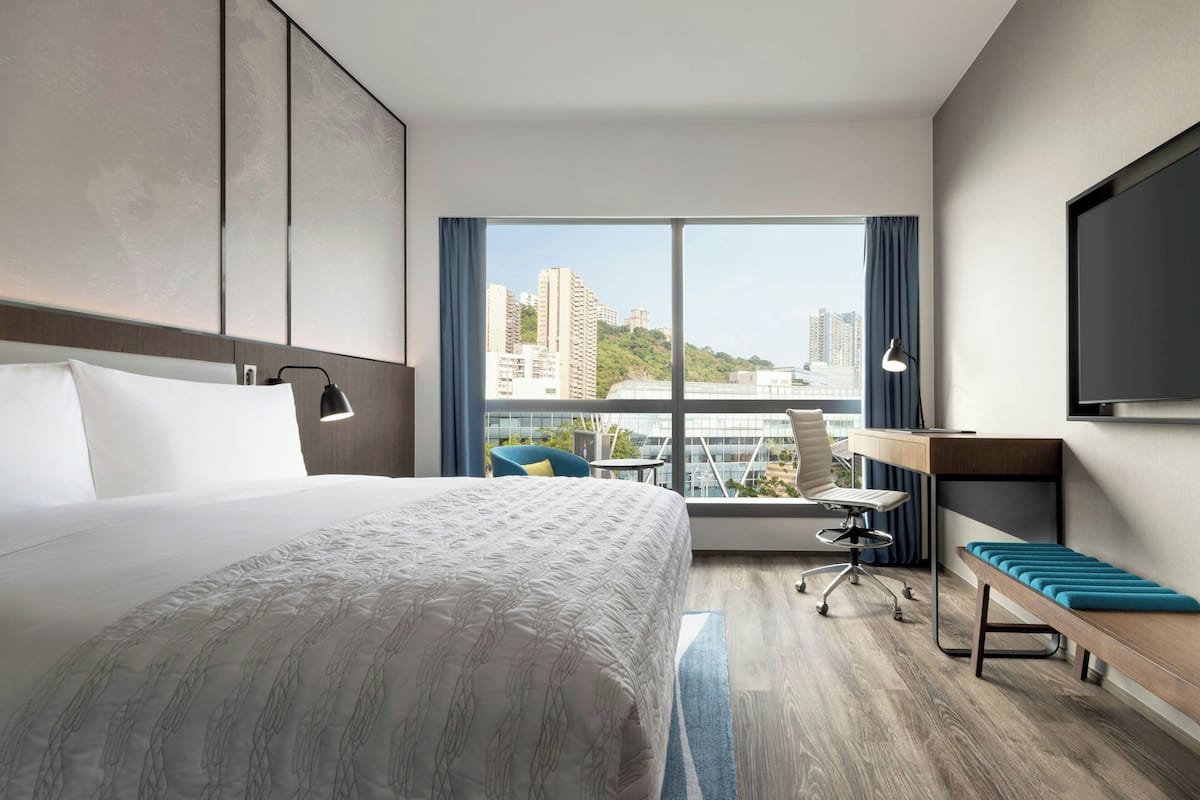 A hotel room with a large bed, a desk, a chair, a TV, and a view of the city.
