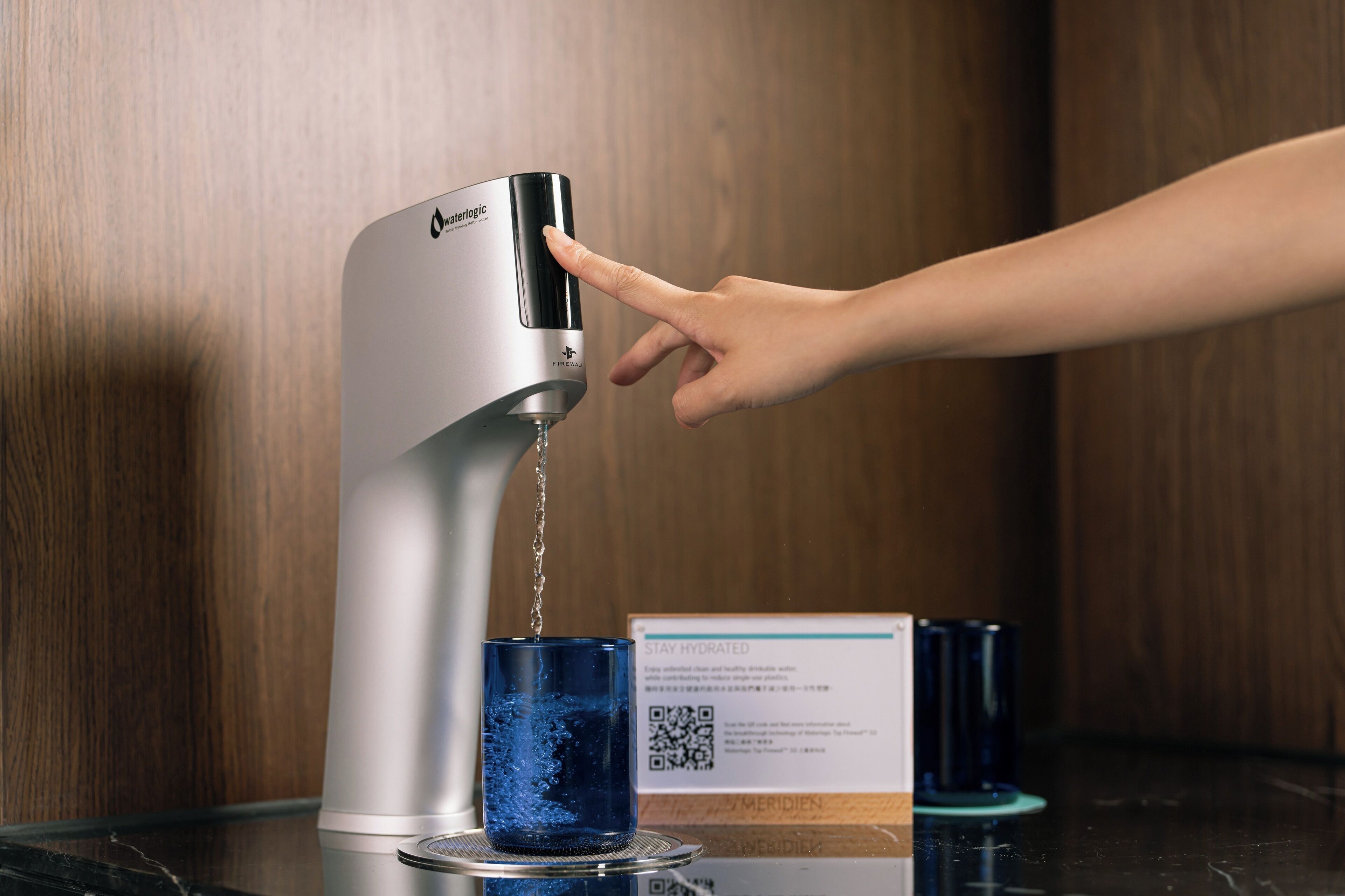 A hand pressing a water dispenser, a glass of water, and a QR code on a counter.