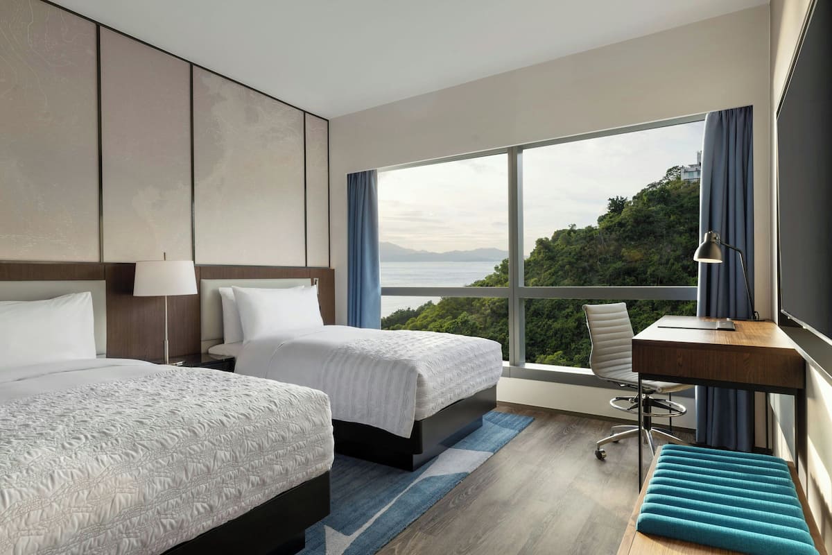 A modern hotel room with two beds, a desk, and a large window offering a view of the sea and hills.