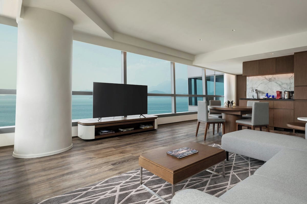 A modern living room with a large flat-screen TV, a sofa, a coffee table, and a dining area with a view of the sea.