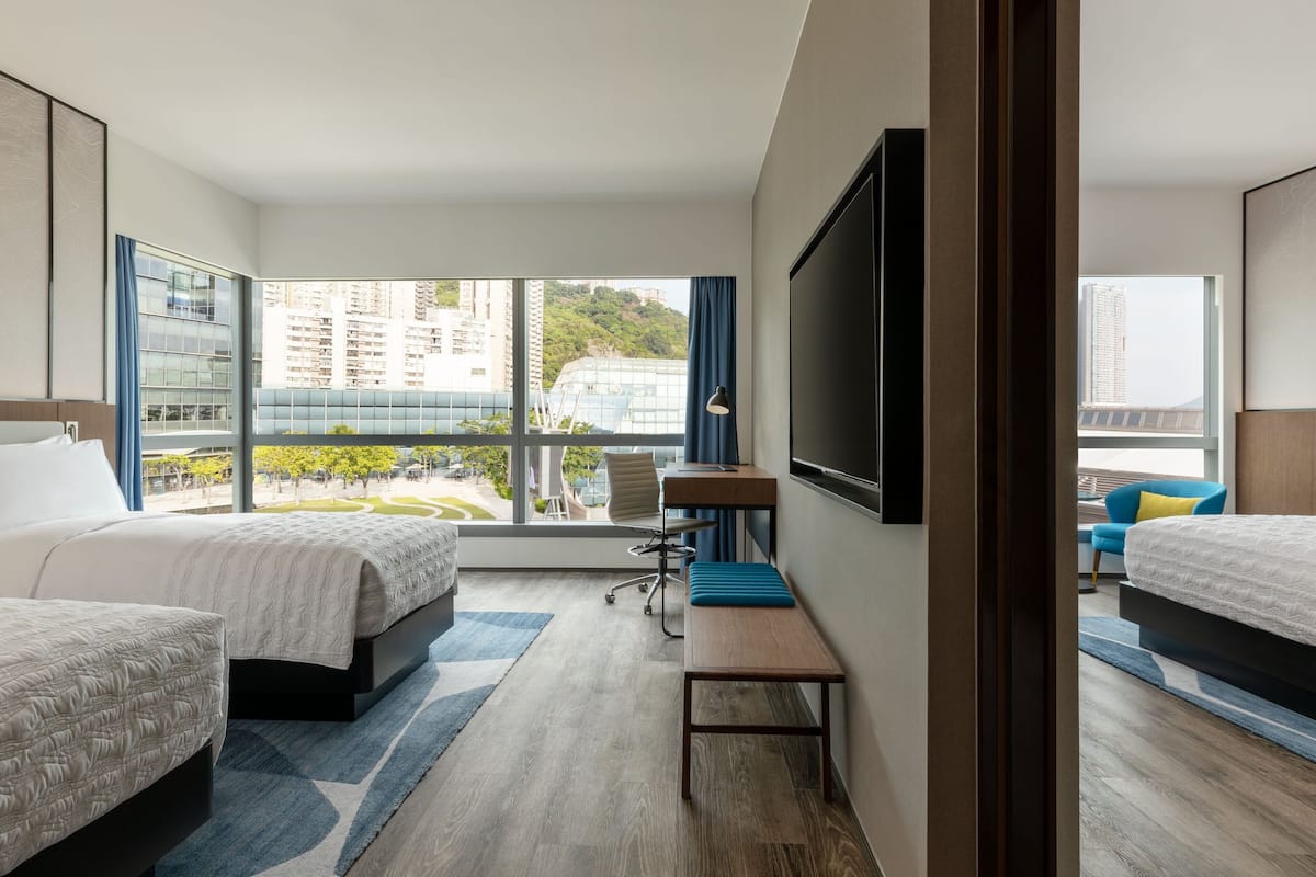 A hotel room with two beds, a desk, a chair, a TV, and large windows offering a city view.