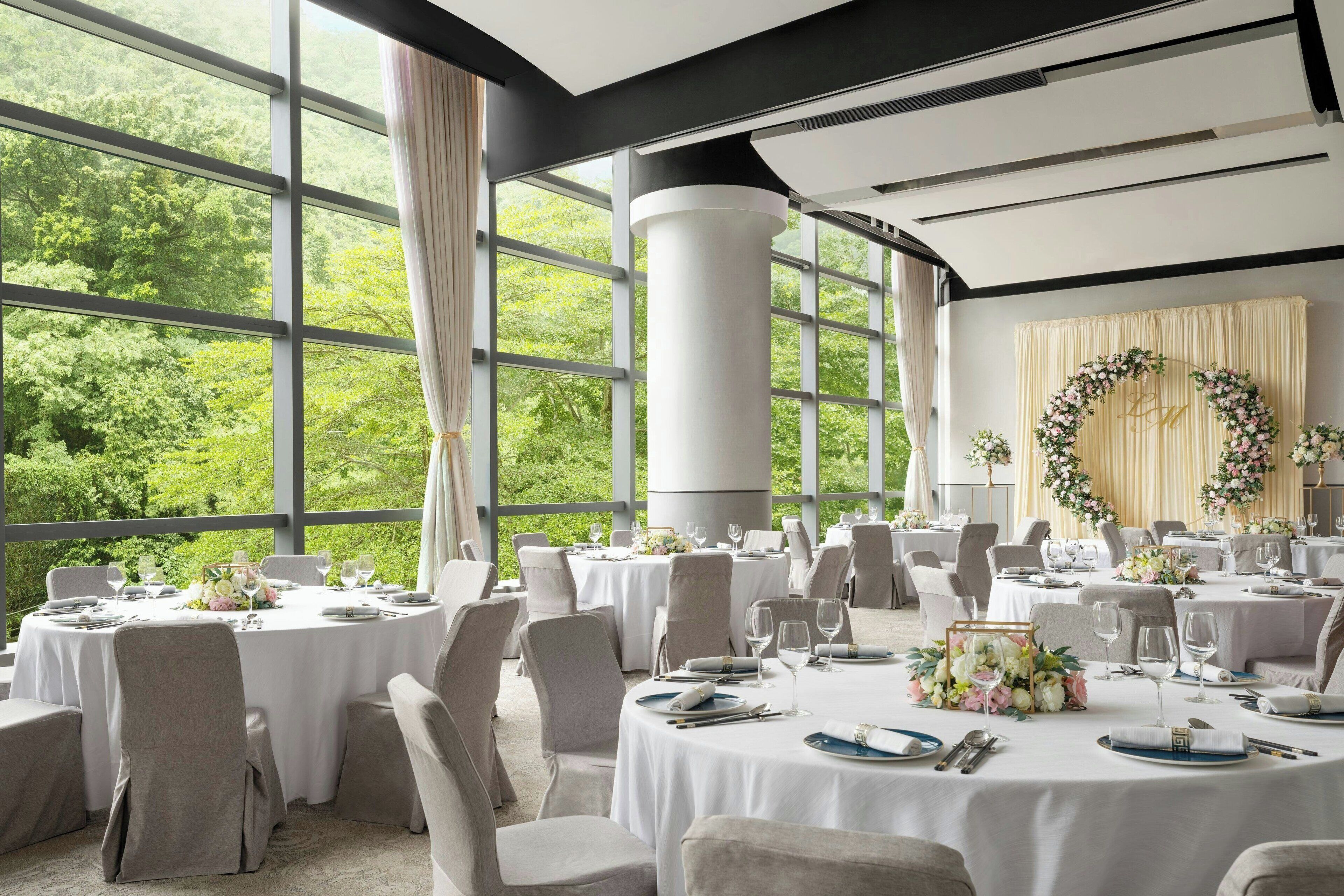 A banquet hall with round tables set for dining, large windows with a view of greenery, and a decorative wreath on the wall.