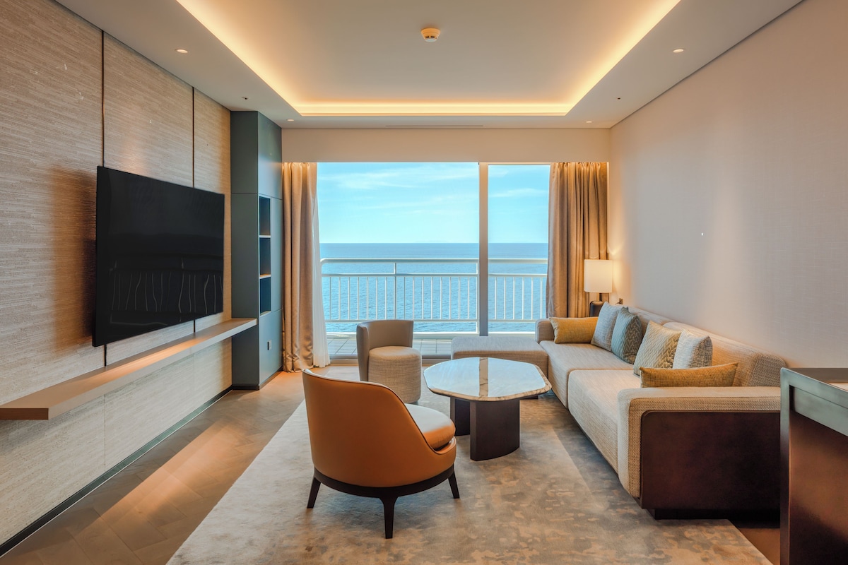 A modern hotel room with a large TV, a sofa, a chair, a coffee table, and a view of the sea.