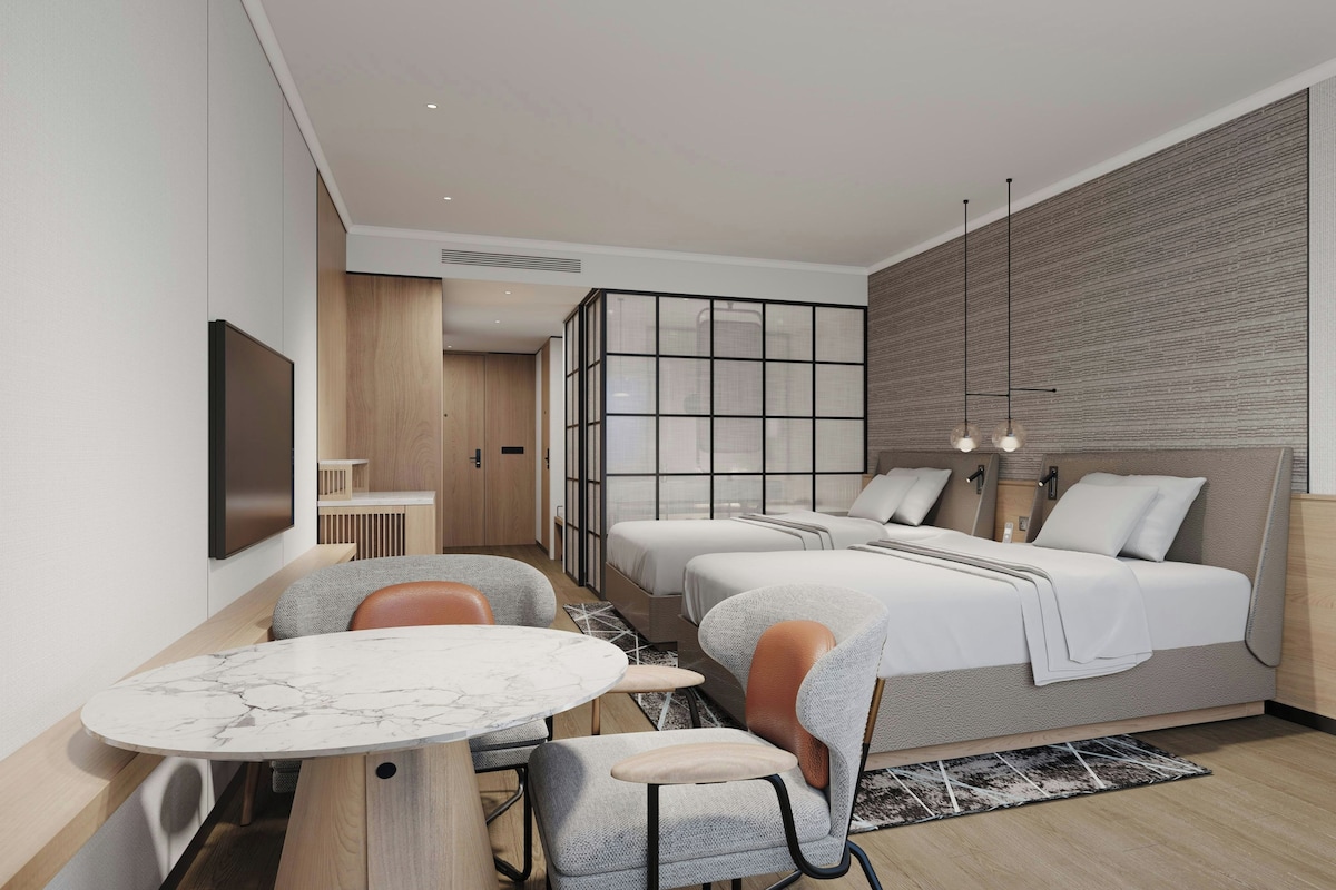 Una habitación de hotel moderna con cama, dos mesitas de noche, un rincón de descanso con sillas, televisión y un ventanal grande.