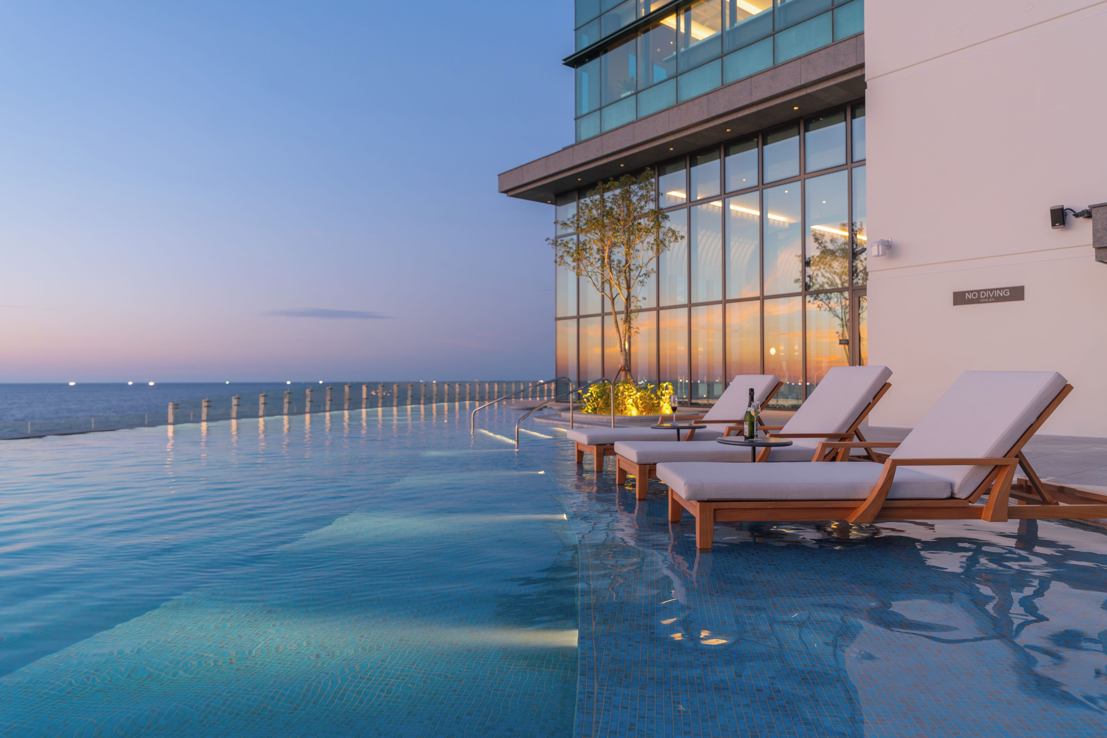 Una piscina infinita con sillones de descanso, una pared de vidrio y vista al mar.