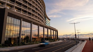 A modern hotel with a curved facade, large windows, and a landscaped area with a road and pedestrian path.