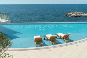 A pool with the word 'SHERATON Jeju' painted on the surface, surrounded by lounge chairs and a view of the sea.