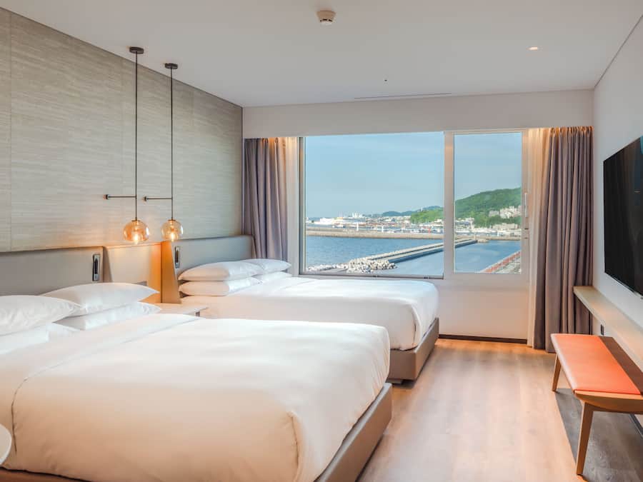 A hotel room with two beds, a TV, a desk, and a view of a harbor.