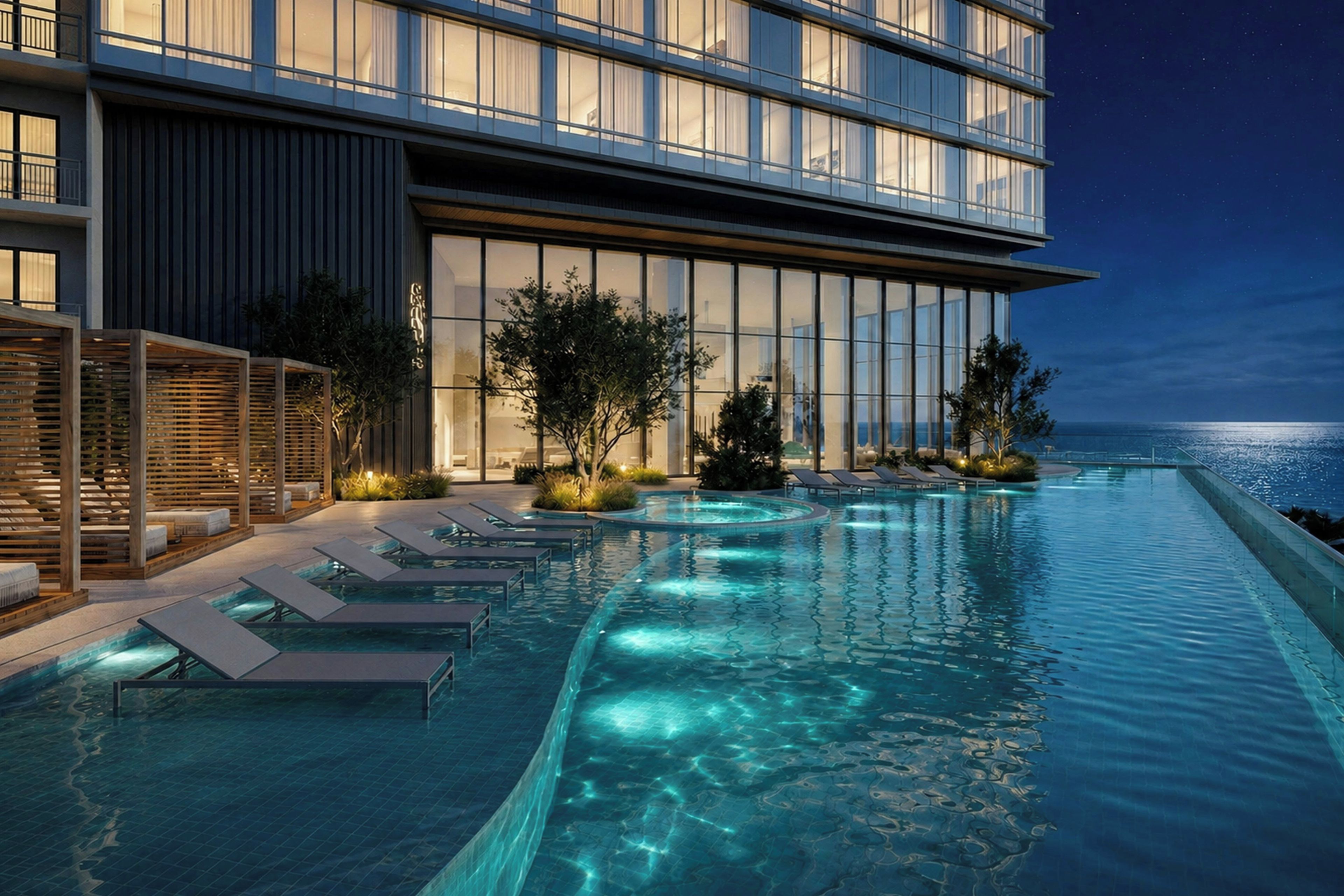 A modern hotel with a poolside area, lounge chairs, and a view of the ocean at night.