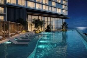 A modern hotel with a poolside area, lounge chairs, and a view of the ocean at night.
