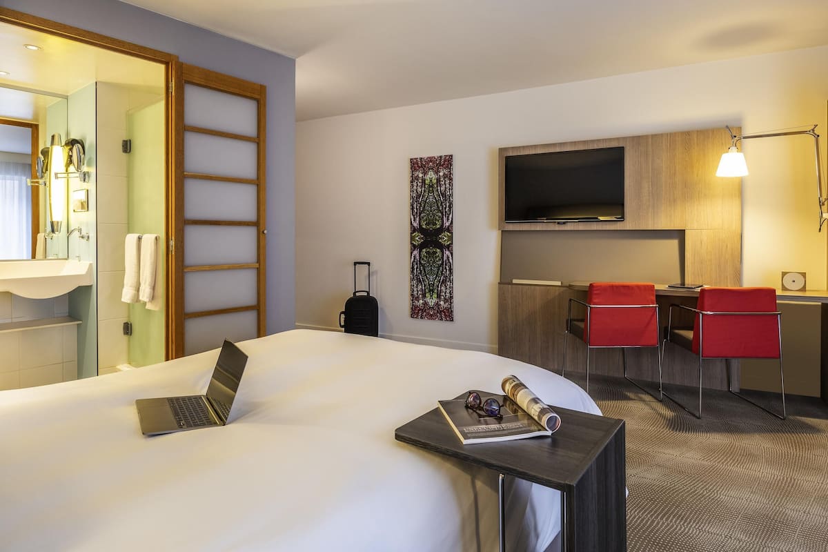 A hotel room with a bed, a desk with a laptop, a TV, and a red chair.