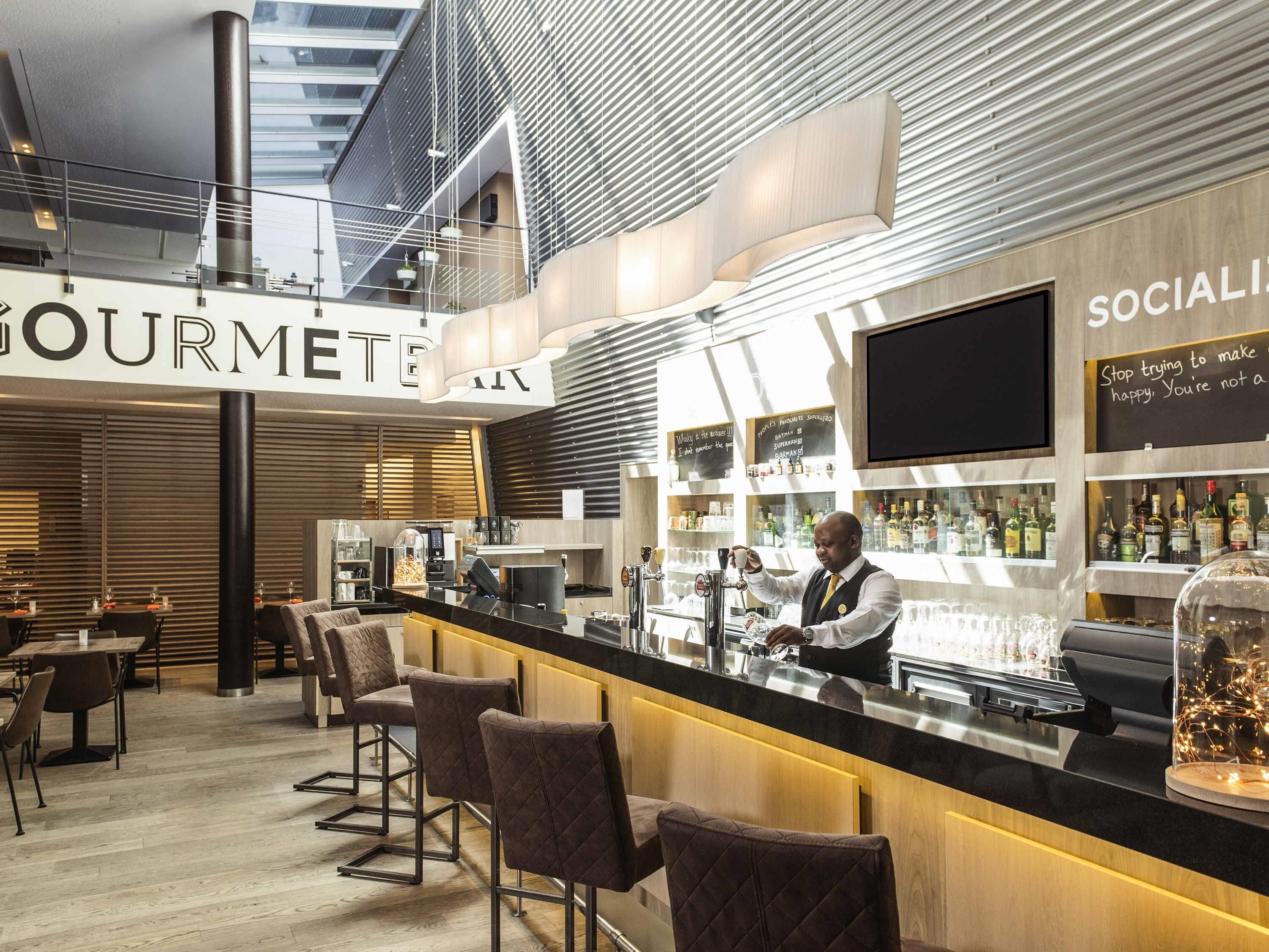 A modern bar area with a high ceiling, bar stools, a counter with a bartender, and shelves with bottles.