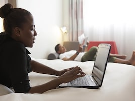 Una persona usando una computadora portátil en una cama de habitación de hotel, con otra persona acostada al fondo leyendo un libro.