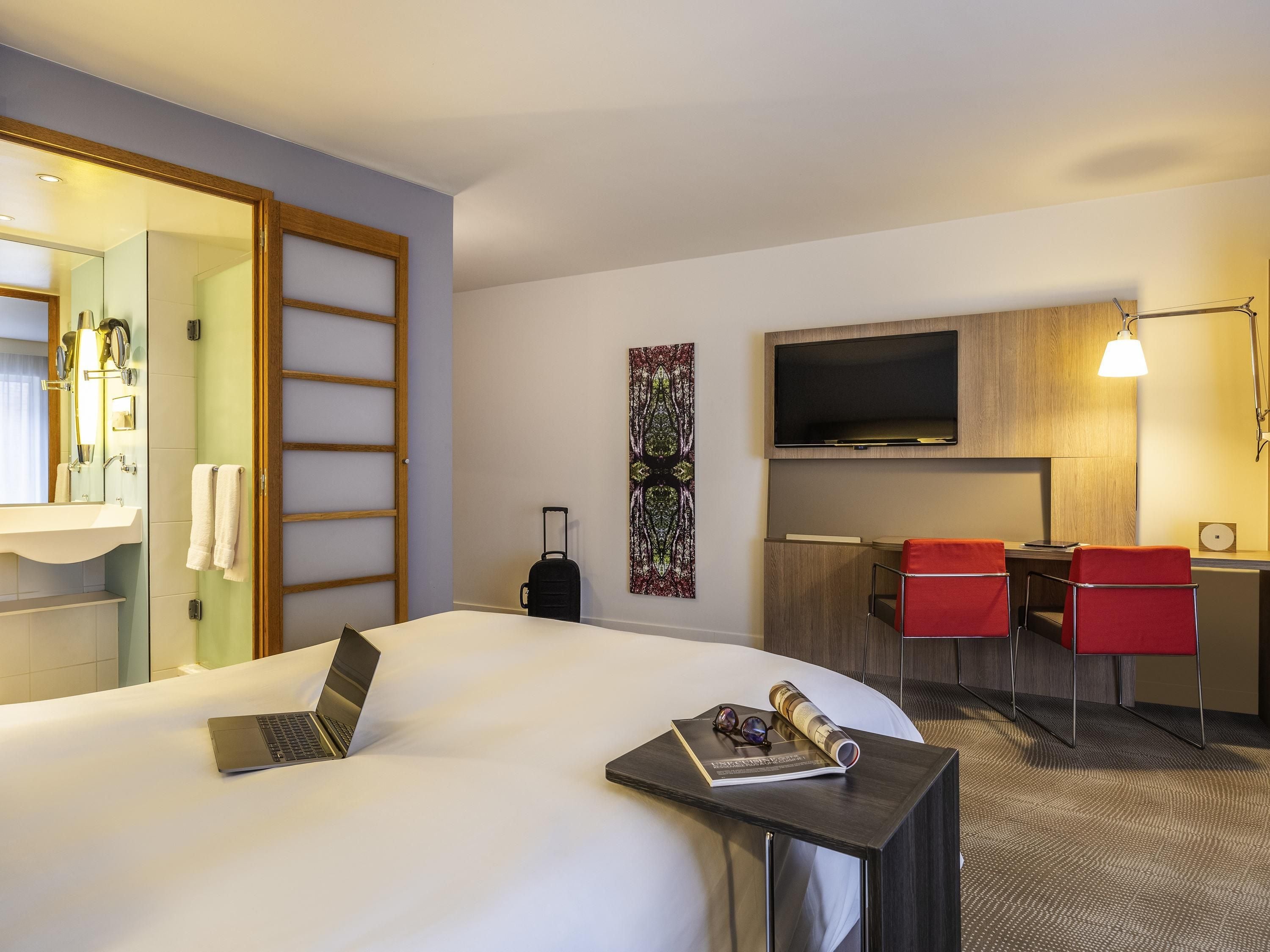 Een hotelkamer met een bed, een bureau met een laptop, een televisie en een rode stoel.