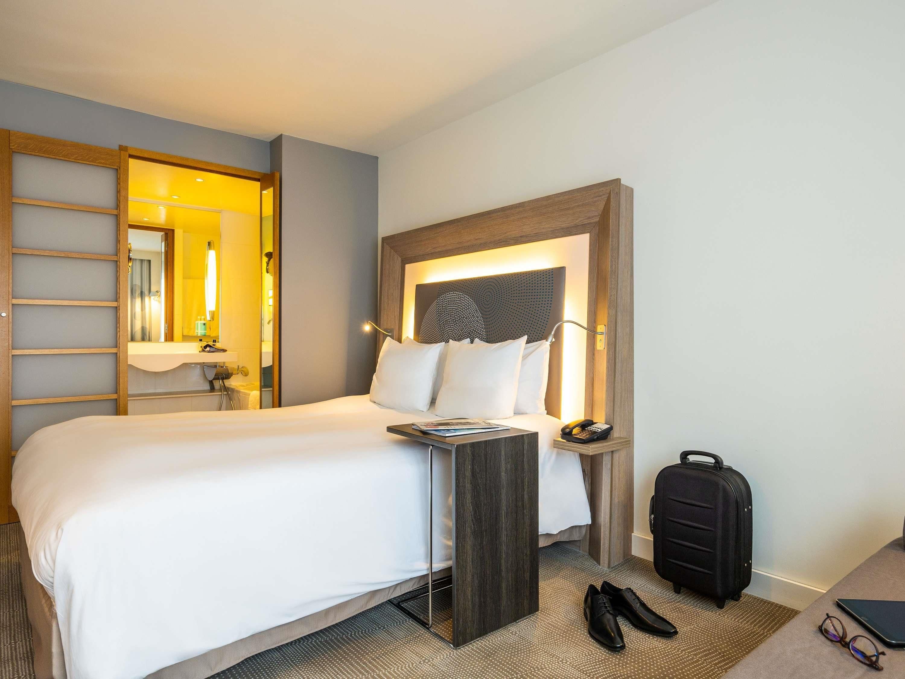 A hotel room with a bed, bedside table, lamp, phone, suitcase, and a pair of shoes.