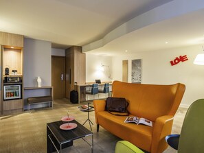 Habitación de hotel con sofá, una mesita con una revista, un banco, kitchenette y un escritorio con sillas.