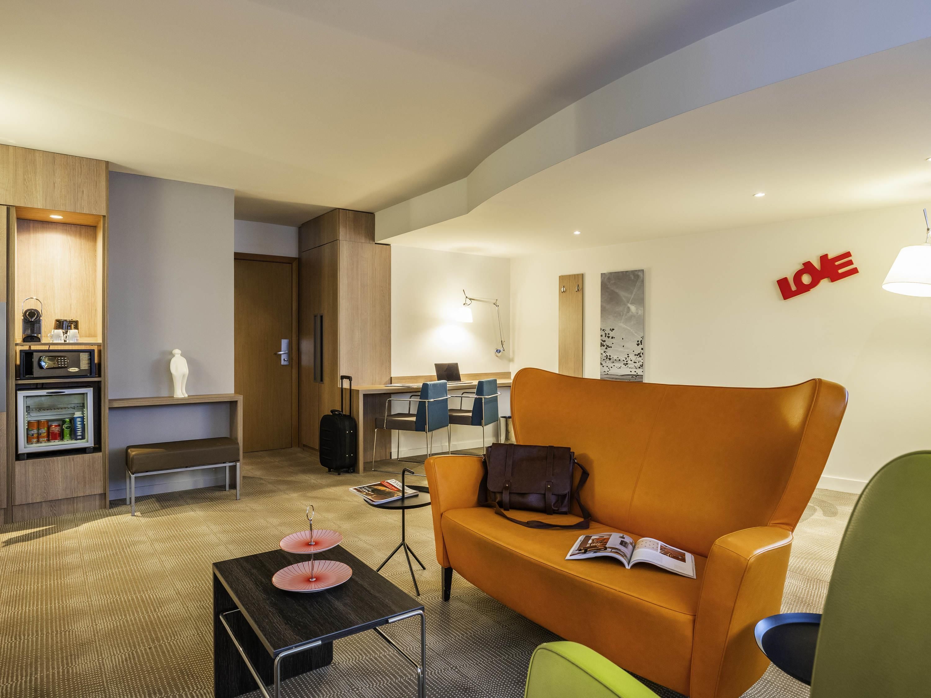 A hotel room with a sofa, a small table with a magazine, a bench, a kitchenette, and a desk with chairs.