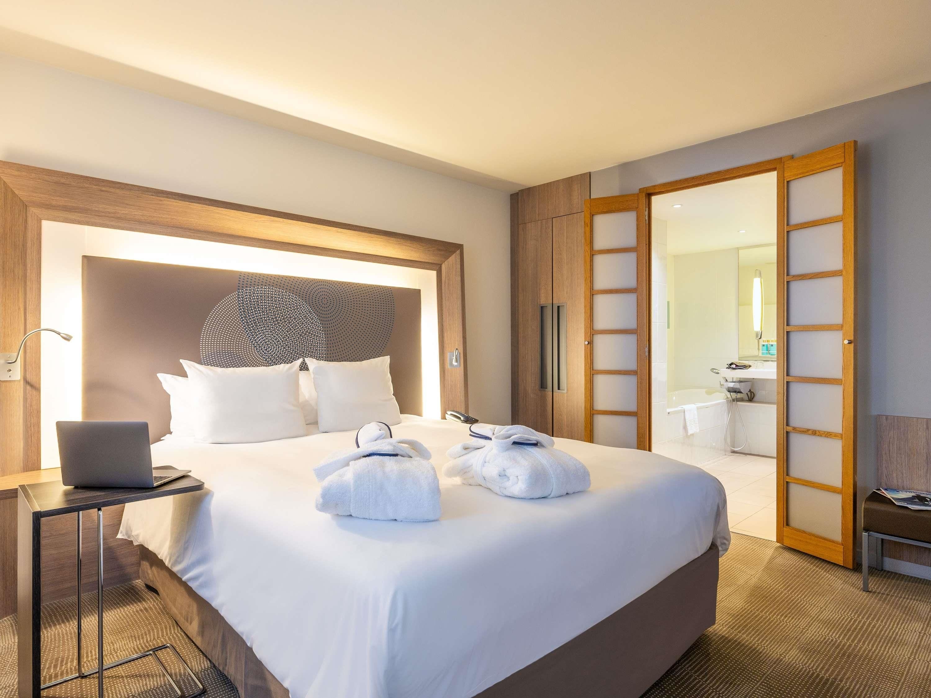 A hotel room with a large bed, a laptop on a bedside table, and a bathroom visible through an open door.