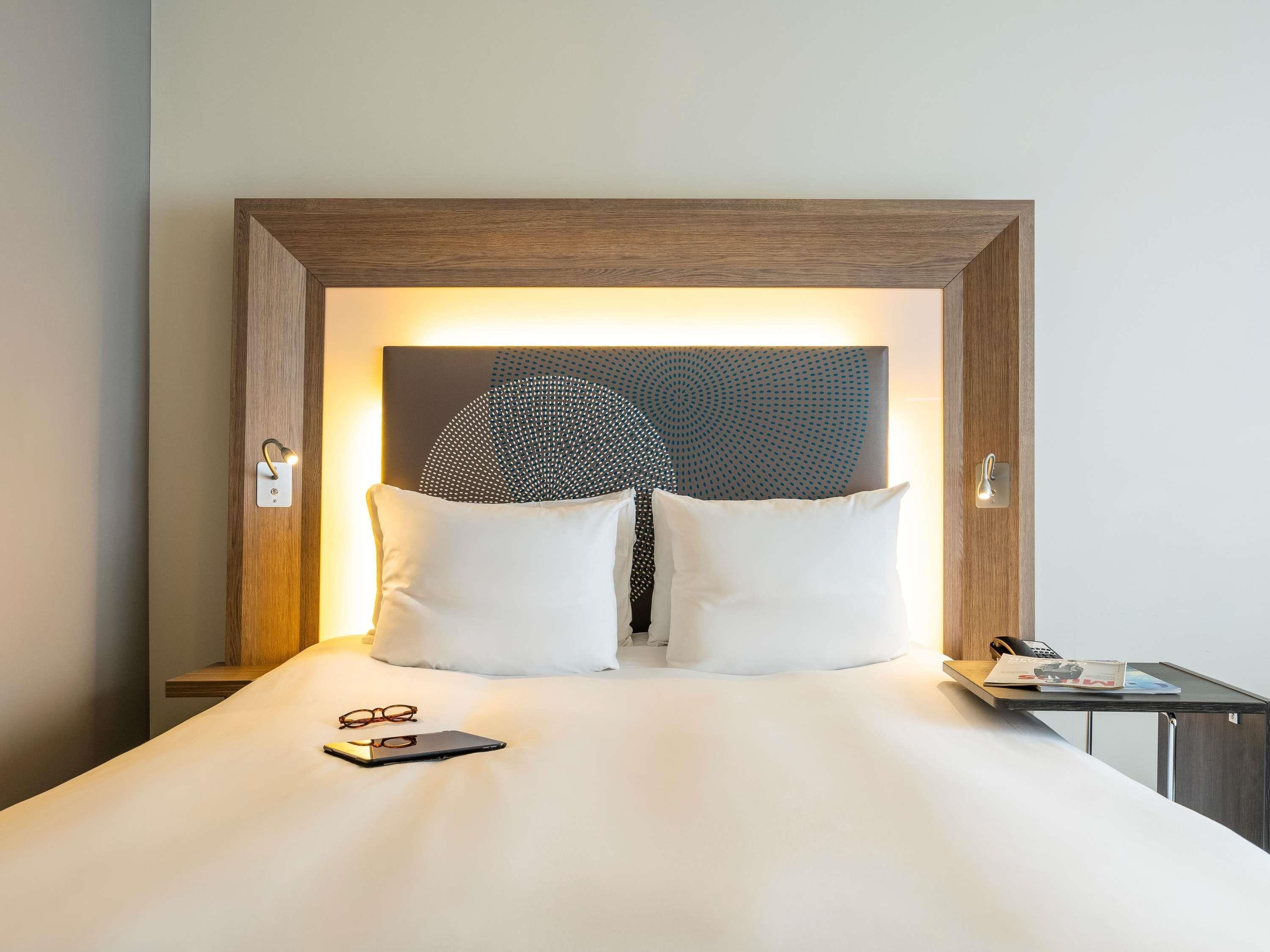 A hotel room with a large bed, two pillows, a bedside table with a magazine, and a wall-mounted light fixture.