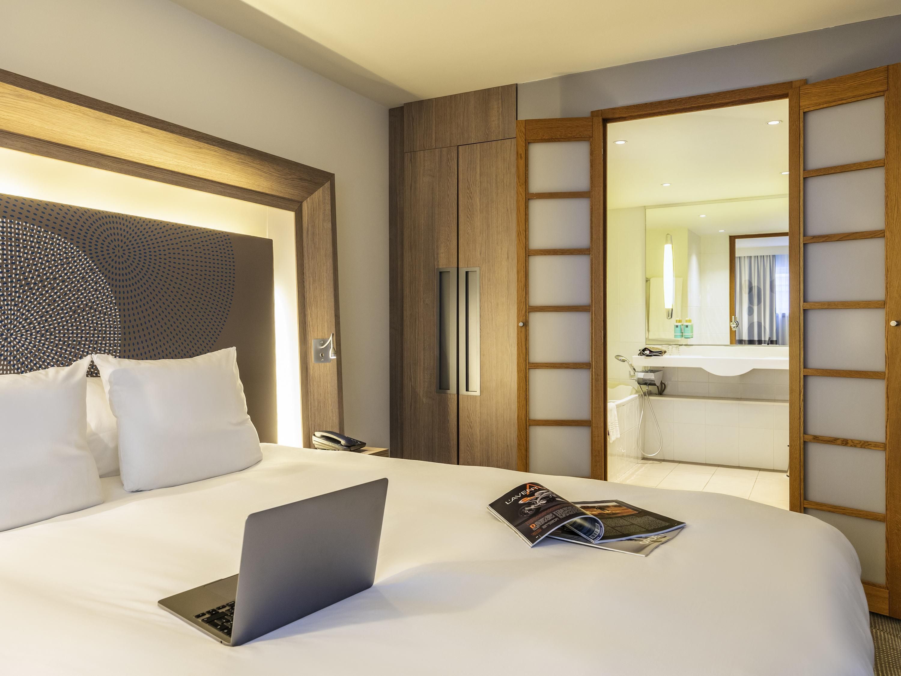 Hotelkamer met een bed, een laptop, een tijdschrift, een telefoon en een zichtbare badkamer door een open deur.