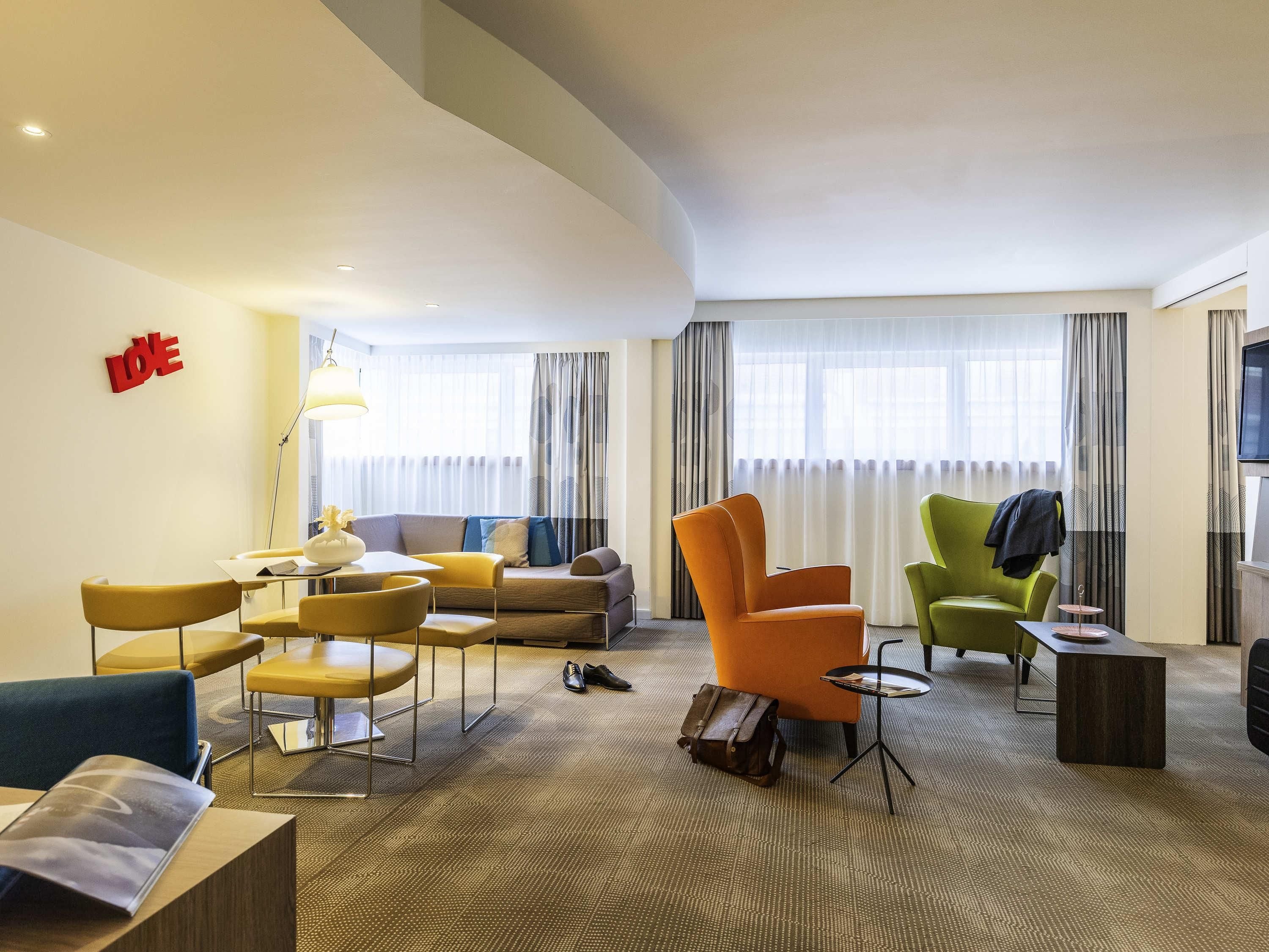 A hotel room with a sofa, armchairs, a small table, a lamp, and a TV.