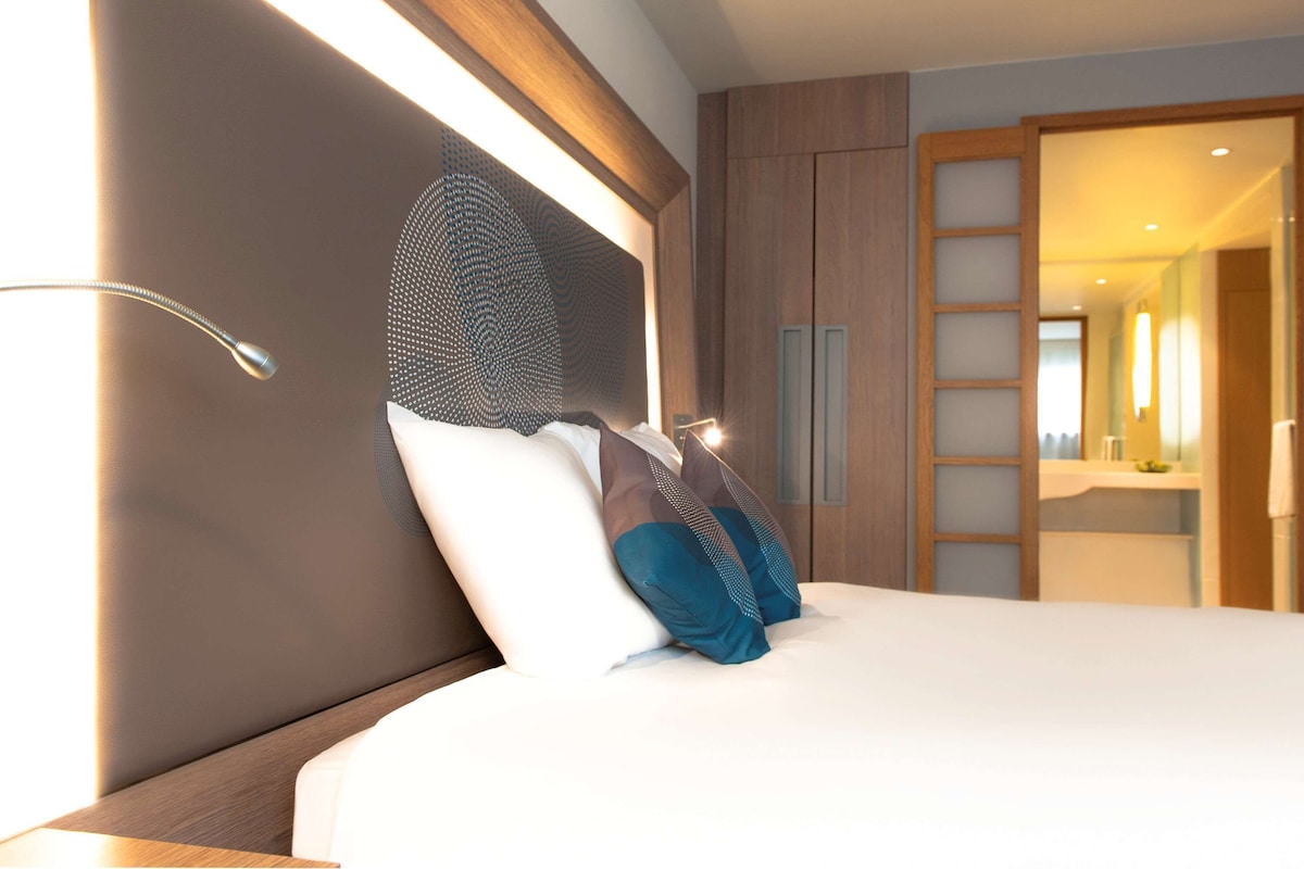 Une chambre d’hôtel moderne avec un grand lit, une tête de lit ornée d’un motif géométrique et une armoire en bois.