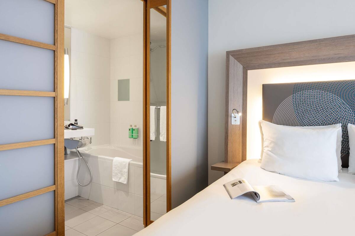 A hotel room with a large bed, a bedside table with a book, a shower area with a bathtub, and a wooden-framed mirror.