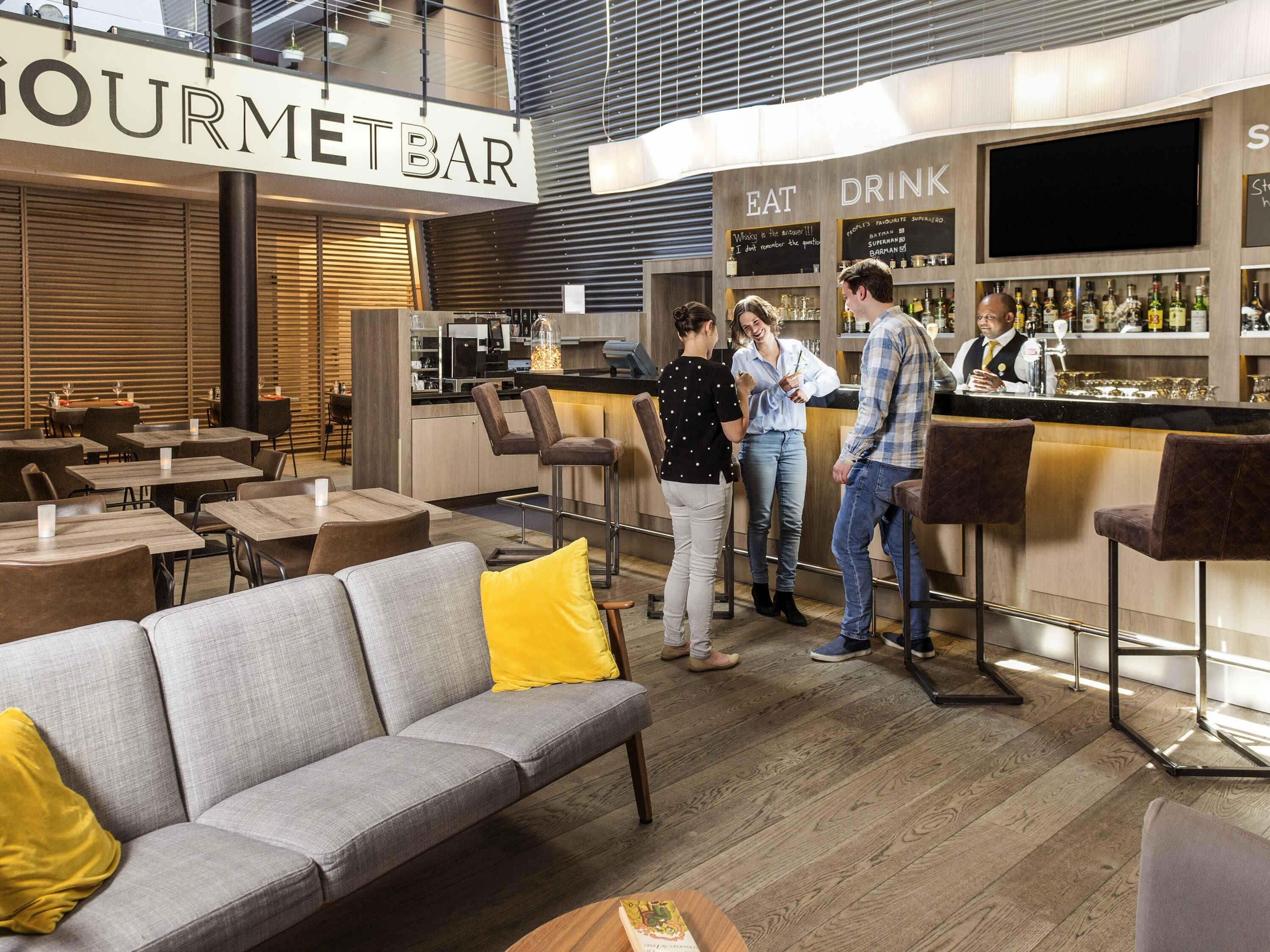 A modern bar area with a sign reading "GOURMET BAR," a counter with a bartender, and seating arrangements including a grey sofa and bar stools.