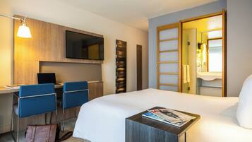 Quarto de hotel com cama, escrivaninha com cadeira, televisão e banheiro visível por uma porta aberta.