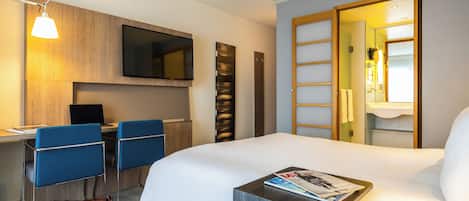 Habitación de hotel con una cama, un escritorio con silla, un televisor y un baño a la vista a través de una puerta abierta.