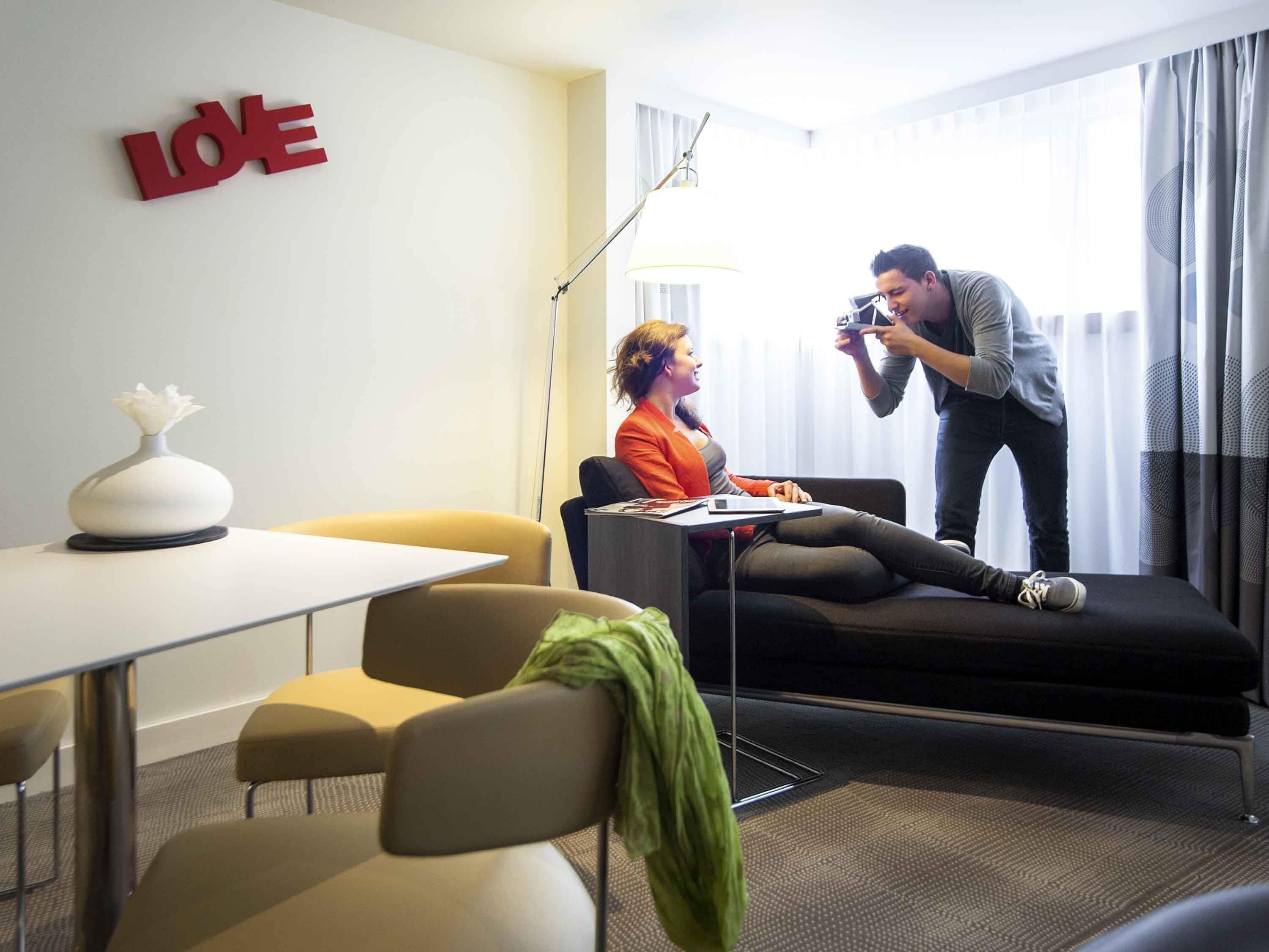 Une chambre d’hôtel avec un canapé, une table sur laquelle est posé un vase, et un panneau « LOE » accroché au mur.