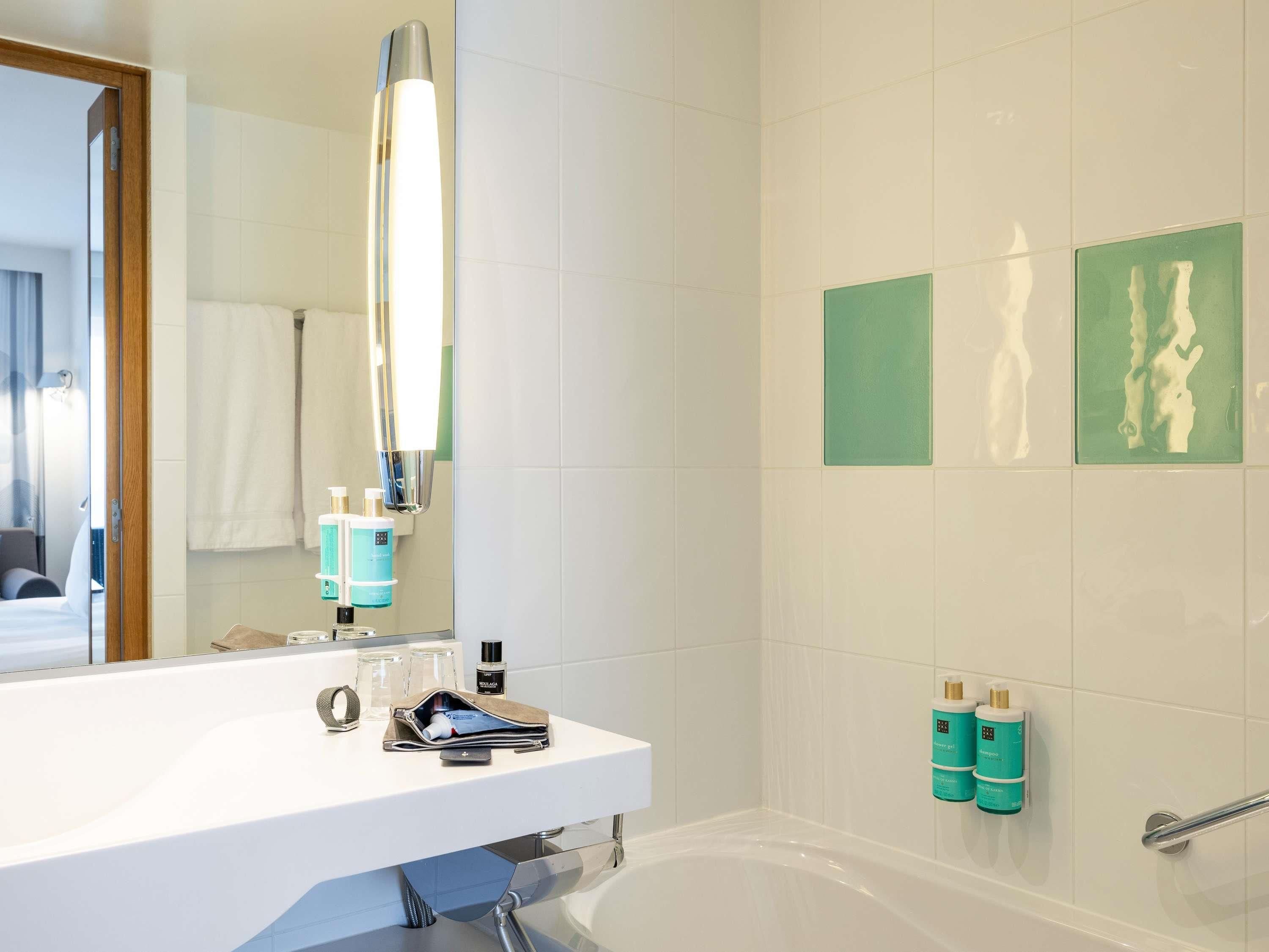 Une salle de bain moderne avec un lavabo blanc, un miroir et une baignoire. On trouve des articles de toilette sur le lavabo et un porte-serviettes au-dessus du miroir.