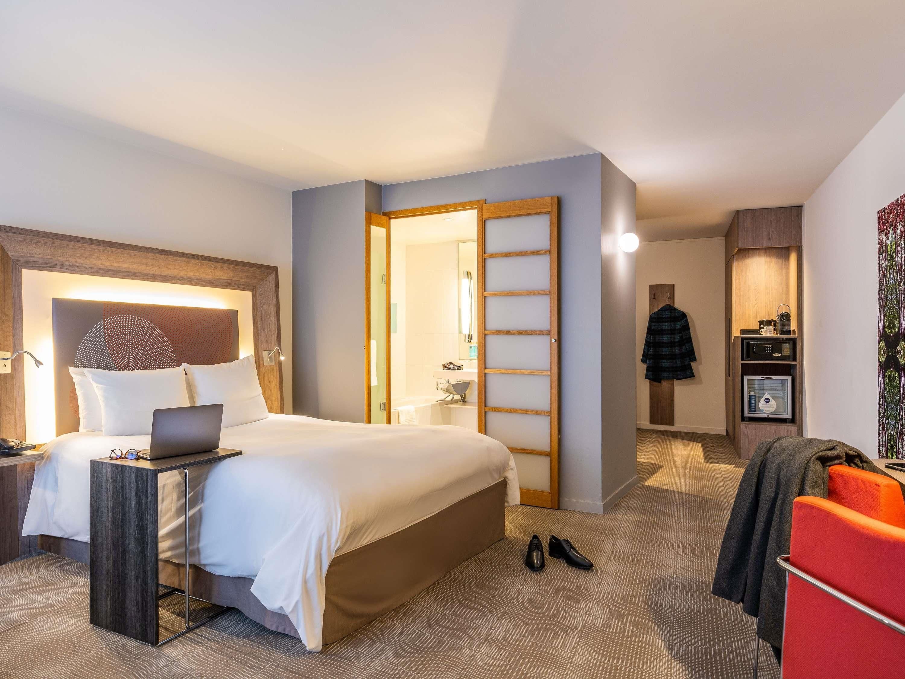 Habitación de hotel con cama, escritorio, ordenador portátil, zapatero y perchero.