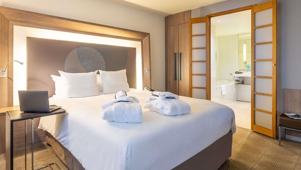 Une chambre d’hôtel avec un grand lit, un ordinateur portable posé sur une table de chevet, et une salle de bain visible à travers une porte ouverte.
