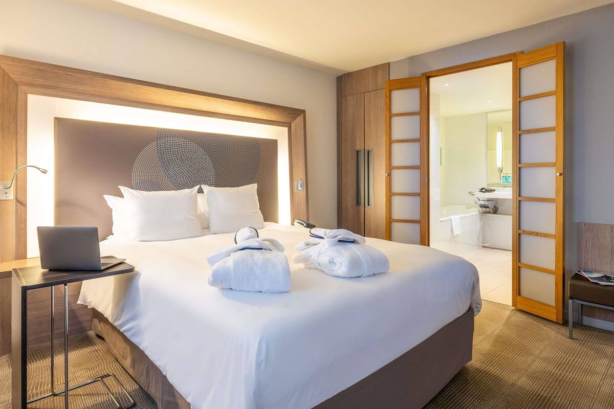 A hotel room with a large bed, a laptop on a bedside table, and a bathroom visible through an open door.
