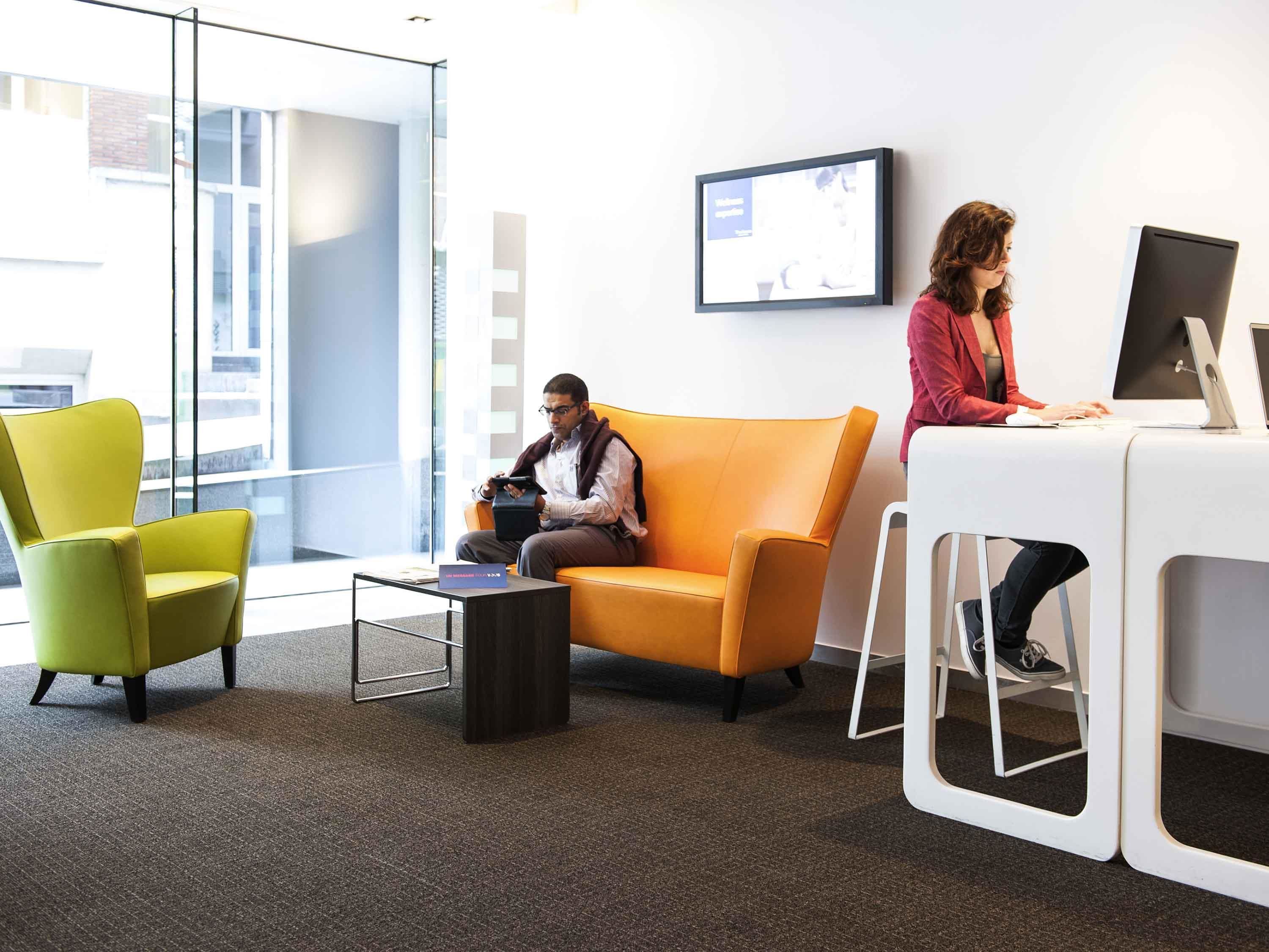 Un espace de bureau moderne avec un mur de verre, un canapé jaune, un fauteuil vert et un bureau blanc équipé d’un ordinateur.