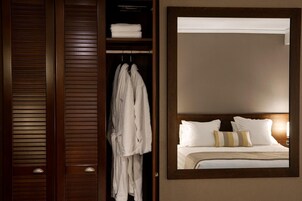 Quarto de hotel com guarda-roupa de madeira, um espelho grande, cama com roupa de cama branca e almofadas listradas, e mesa de cabeceira com uma luminária.