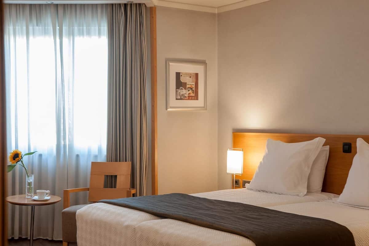 A hotel room with a bed, a chair, a table with a cup and a vase of flowers, and a picture on the wall.